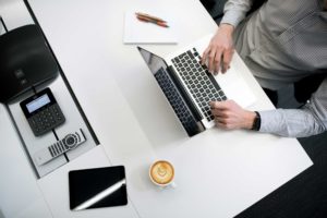 Vitt skrivbord ovanifrån, där en man skriver på en laptop med en kopp kaffe bredvid sig.