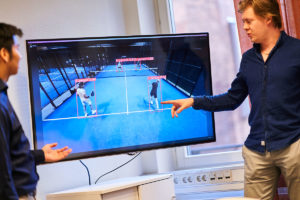 Två personer står framför en skärm som visar en bild från en padel-match. På skärmen syns data över hur spelarna rört sig och presterat under matchen.