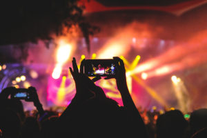 En person står i ett publikhav och håller upp en telefon som fotar en scen. På scenen syns strålkastare i olika färger.