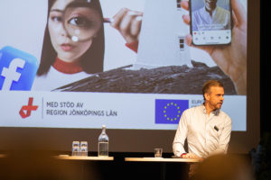 Anders Nyberg står på scen framför en skärm som visar omslagsbilden för en trendrapport. På omslaget syns en bild av en kvinna med ett förstoringsglas i handen samt texten "Med stöd av Region Jönköpings län" och logotypen för Europeiska regionala utvecklingsfonden.