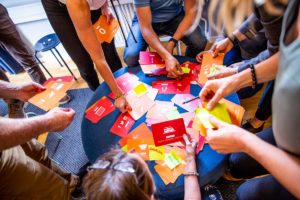 Bilden visar händer som håller i olika färgglada kort som ligger på ett litet bord. Personerna ser ut att hålla på med ett spel eller en workshop.