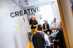 En grupp personer som håller en workshop är fotade genom en glasruta med texten "Creative". En av personerna står framför en vägg med postit-lappar och de andra sitter och diskuterar det som står på väggen.