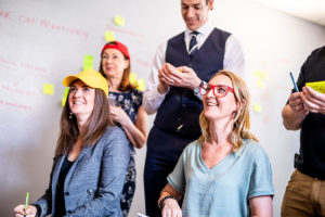 En grupp personer sitter i färgglada kepsar och glasögon och lyssnar på en pitch. De hjälper en entreprenör med pitchträning och att ge feedback på en affärsidé.