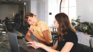 Två glada kvinnor sitter framför var sin dator. De skrattar och pekar på skärmen.
