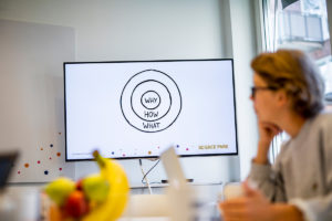 En skärm visar föreläsaren Simon Sineks modell "The Golden Circle". Modellen består av tre cirklar med texten "why" i mitten. I förgrunden syns silhuetten av en korthårig person med glasögon.