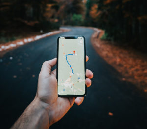 Närbild på en hand som håller i en telefon. På telefonen används en GPS-app. Personen befinner sig på en väg med höstig skog runt omkring sig. 