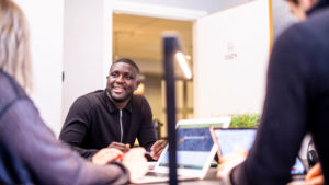 En entreprenör sitter i ett mötesrum. Han är glad och pratar med två personer som syns i förgrunden.