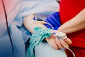 En person ligger på en sjukhusbrits med en mätare kopplade till sitt finger. En person i röda sjukhuskläder står vid sidan och håller i personens arm.
