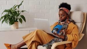 En kvinna sitter i en fåtölj med en laptop i knät. Hon skriver fokuserat. Omgivningen är ljust beige och vit.