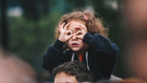 En liten flicka sitter på en vuxen persons axlar. Hon formar händerna till kikare framför ögonen.