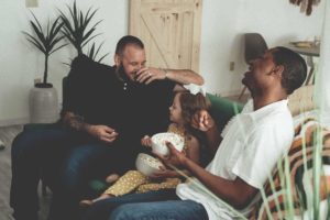 Två pappor sitter i en soffa tillsammans med sin lilla dotter. Flickan håller ett mjukisdjur i famnen och hela familjen skrattar.