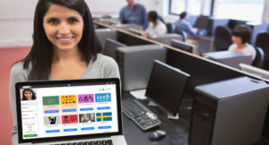 En kvinna står i en lärosal och håller upp en dator som visar lär-plattformen Scoolia på skärmen.