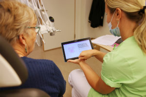 En tandläkare sitter tillsammans med en äldre patient i ett undersökningsrum. tandläkaren visar patienten en app som ska hjälpa henne att borsta tänderna på rätt sätt.