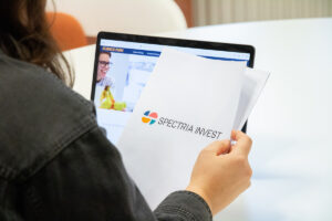 Närbild på en hand som håller i ett pappersdokument med Spectria invests logottyp på. I bakgrunder står en dator på bordet.