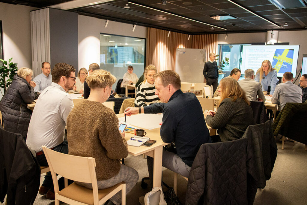 En grupp m&auml;nniskor sitter vid olika bord i ett konferensrum.