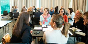 En grupp människor sitter tillsammans vid ett bord och diskuterar. De ser glada ut och i bakgrunden syns en rollup med texten "Nu tar vi sikte på framtiden"