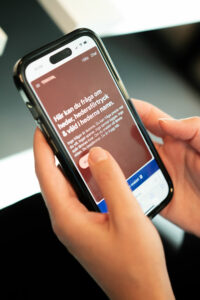 En person håller i en mobiltelefon med Tnkoms chattgränssnitt öppet, där det står att man kan ställa frågor om heder och hedersförtryck anonymt och säkert.