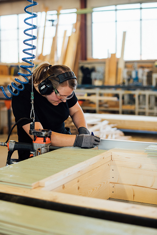 En kvinna står i en fabrik och arbetar med trä. Kvinnan har hörlurar och skyddsglasögon.
